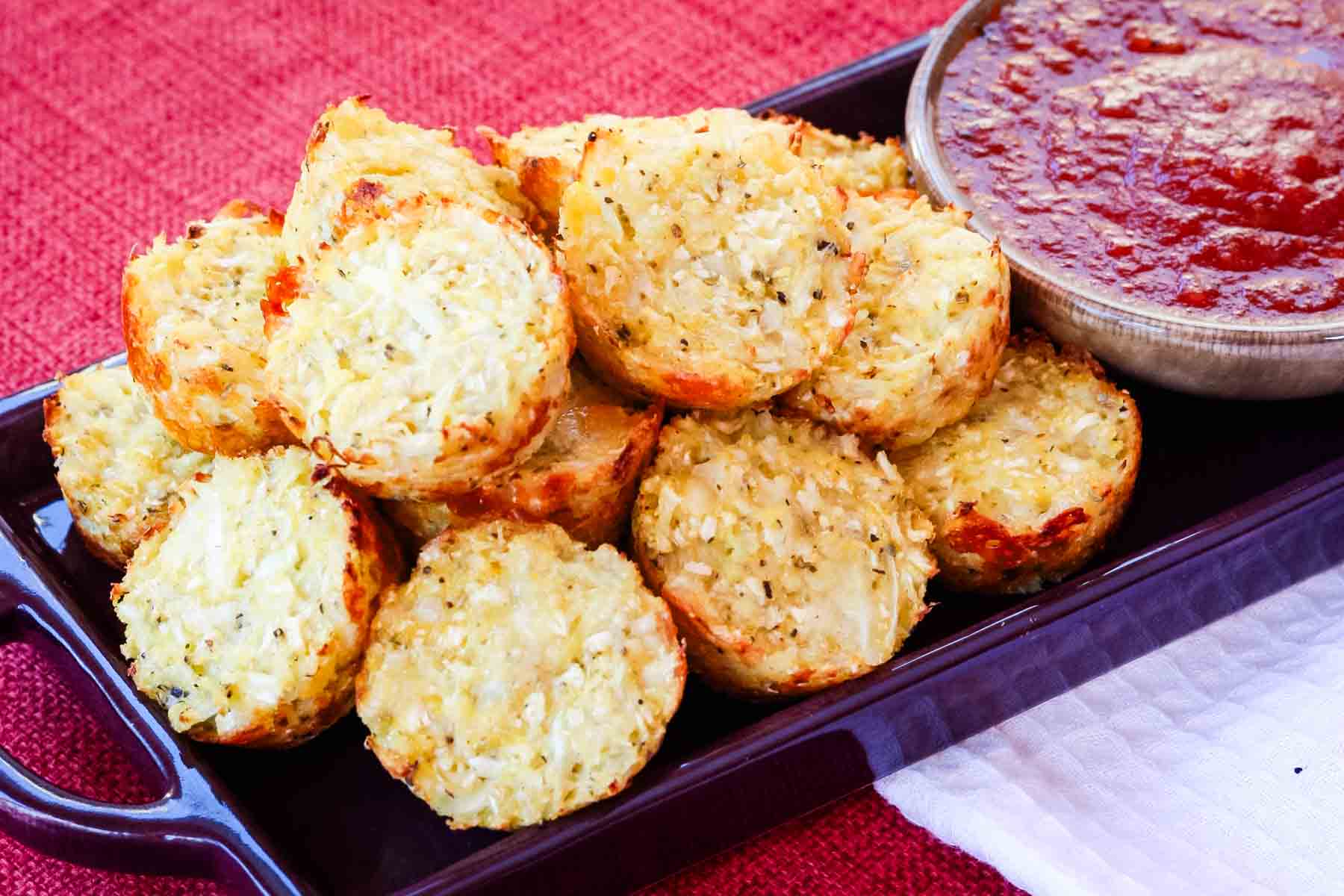 A pile of mozzarella cauliflower tots on a dish with a small bowl of marinara sauce.