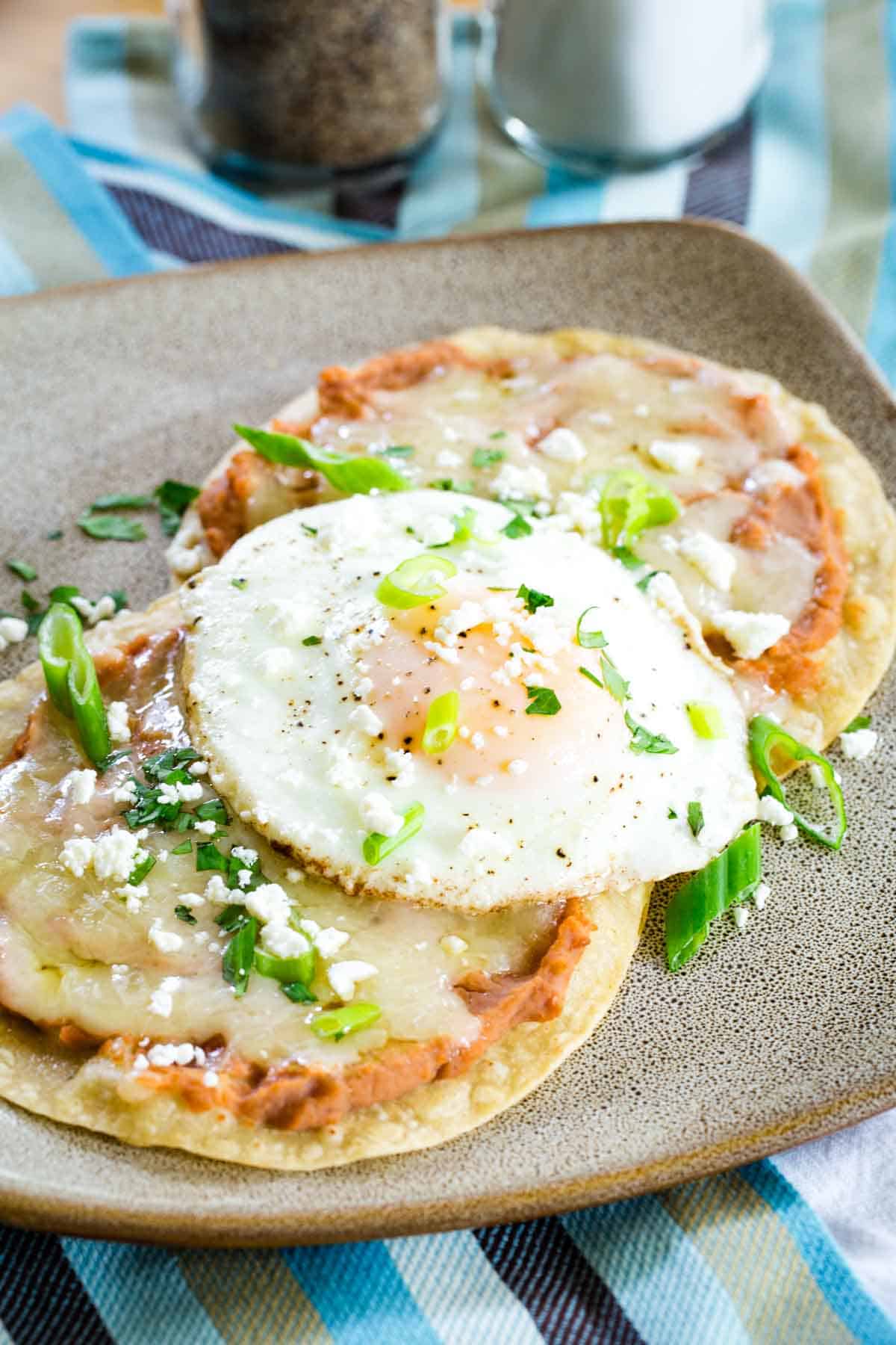 Ready in ten minutes, breakfast tostadas are made with refried beans, melted cheese, and soft fried eggs layered on top of crisp tortillas. Crisp breakfast tostadas are topped with fried eggs.