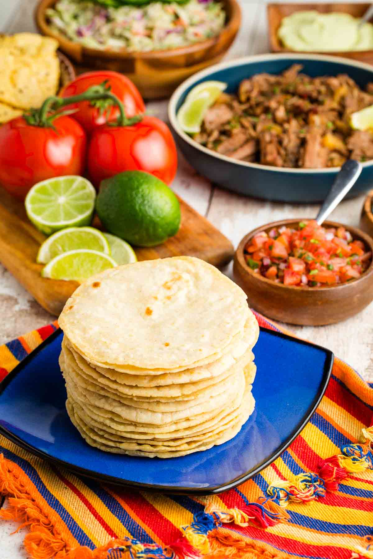 Limes, tomatoes, and bowls of fillings are seen behind a stack of corn tortillas.
