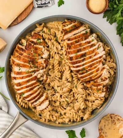 A big pan of garlic parmesan chicken pasta surrounded by cheese and garnishes.