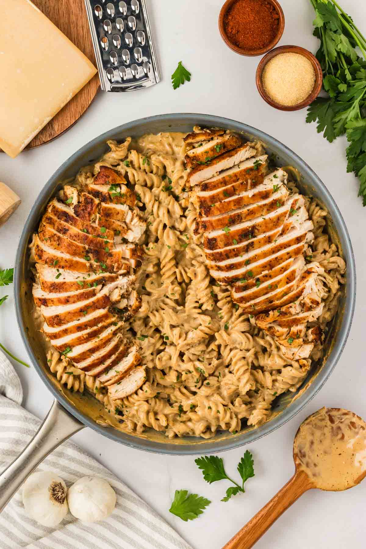 Garlic parmesan chicken pasta drapes tender chicken breasts in a rich and creamy sauce that's paired with gluten free pasta. A big pan of garlic parmesan chicken pasta surrounded by cheese and garnishes.