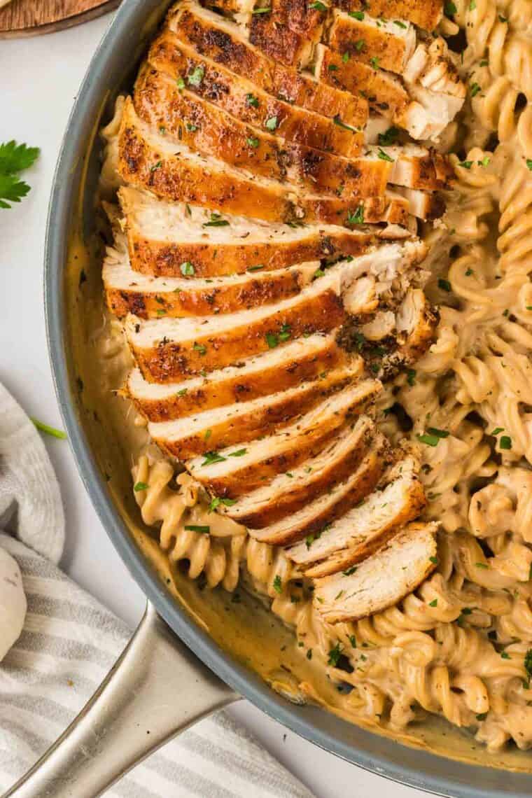 Garlic parmesan chicken pasta drapes tender chicken breasts in a rich and creamy sauce that's paired with gluten free pasta. Slices of chicken are arranged on top of a pan of garlic parmesan chicken pasta.
