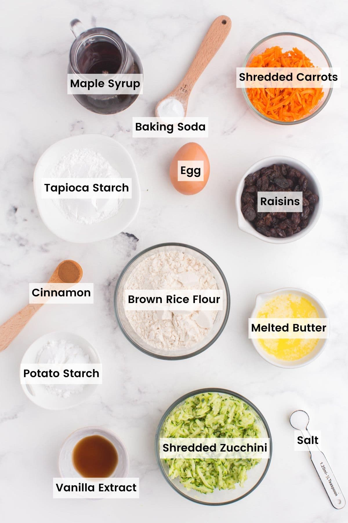 bowls of gluten free flours, shredded carrots and zucchini, vanilla extract, raisins, and melter butter, a pitcher of maple syrup, and egg, and measuring spoons of cinnamon, salt, and baking soda on a marble tabletop with text labels.