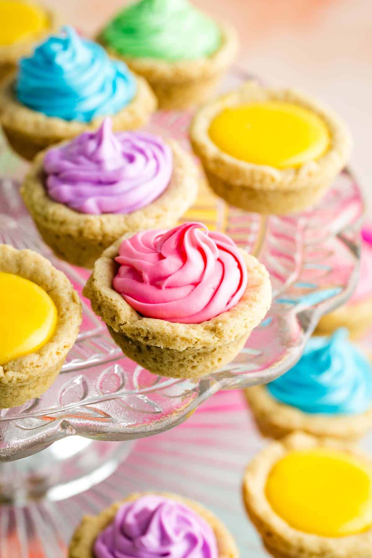 A cake stand holds sugar cookie cups filled with colored frosting.