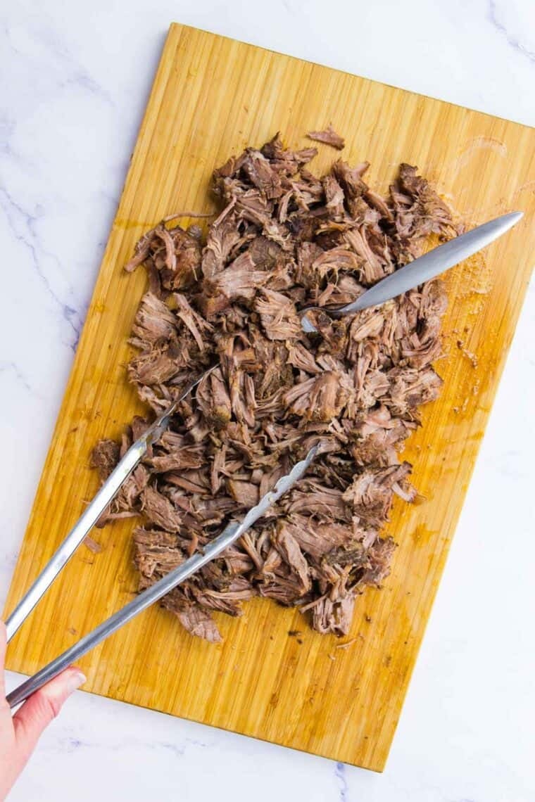 Barbacoa meat is shredded on a cutting board with tongs and a fork.