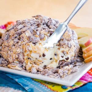 A peanut butter chocolate chip cheese ball covered in Muddy Buddies on a plate with some removed and a small spreader stuck in it.
