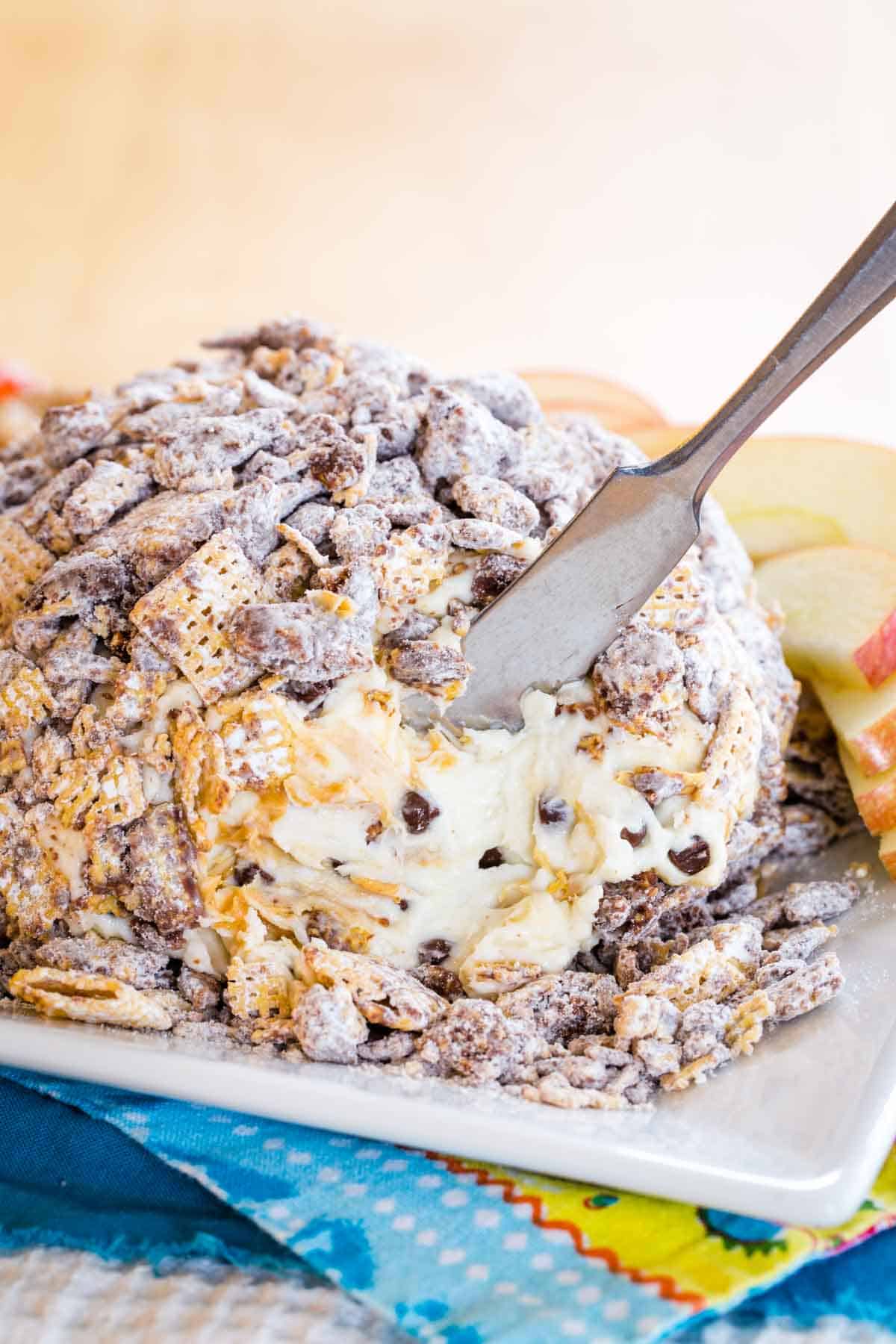 A Muddy Buddy-covered peanut butter chocolate chip cheese ball with some scooped out with a spreader knife.