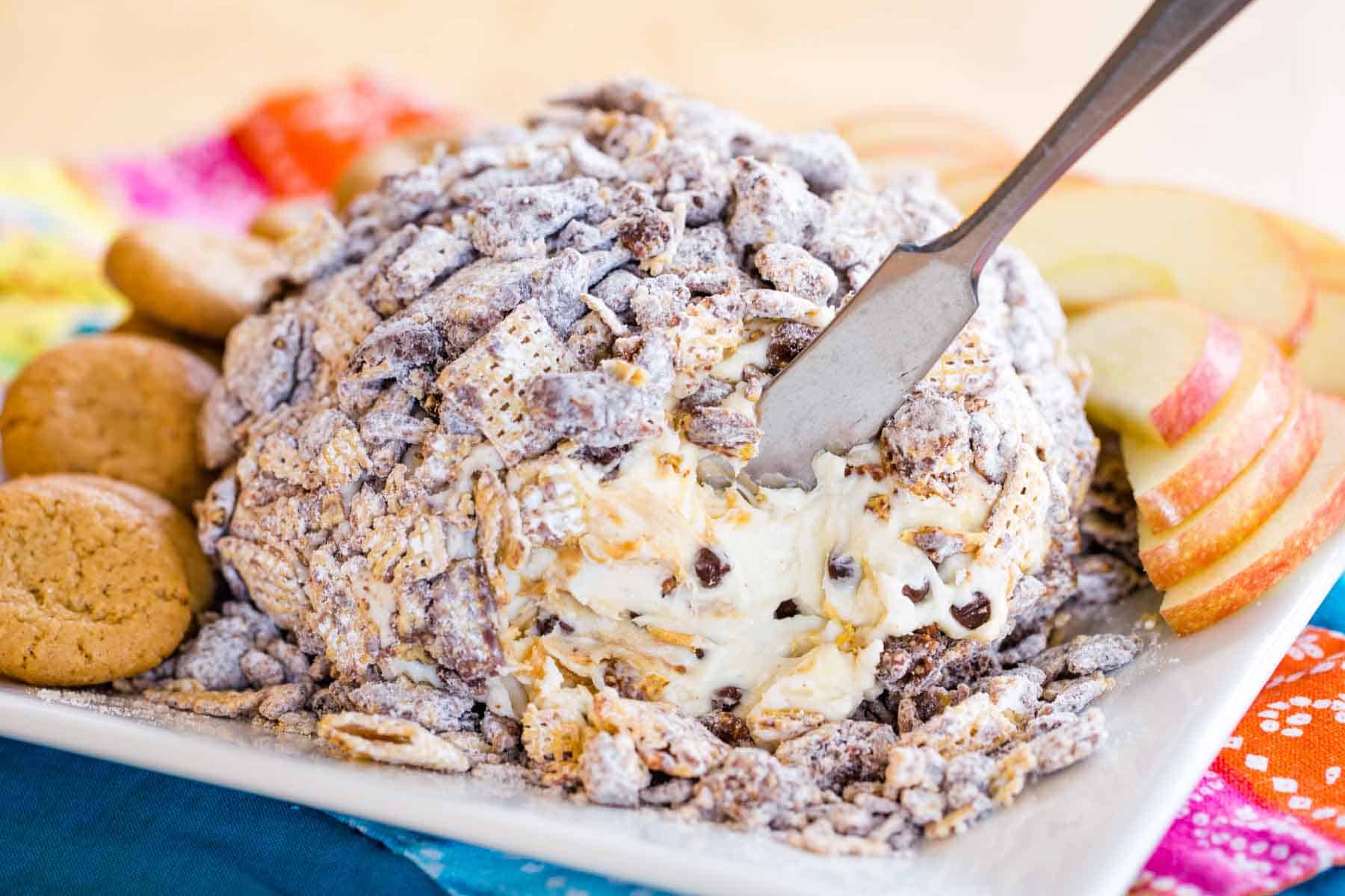 A small spreader knife in a peanut butter chocolate chip cheese ball covered in Muddy Buddies on a plate with apple slices and cookies.