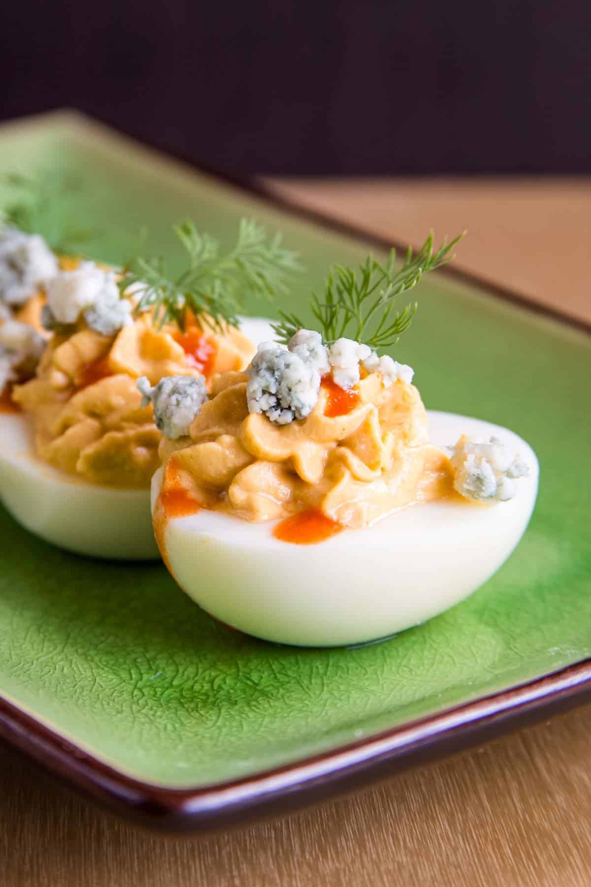 A closeup of one of the buffalo deviled eggs with blue cheese crumbles, a drizzle of hot sauce, and a sprig of dill on toip.