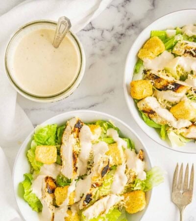 A jar of caesar dressing with a spoon in it and bowls of chicken caesar salad.