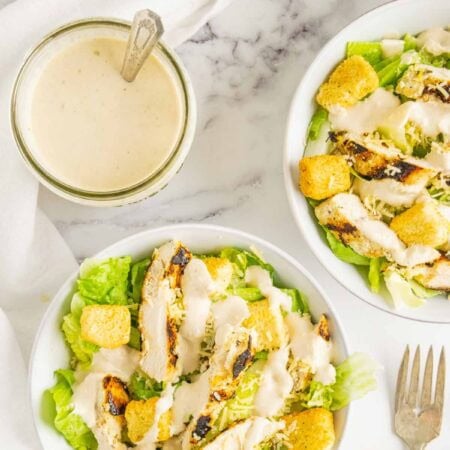 A jar of caesar dressing with a spoon in it and bowls of chicken caesar salad.