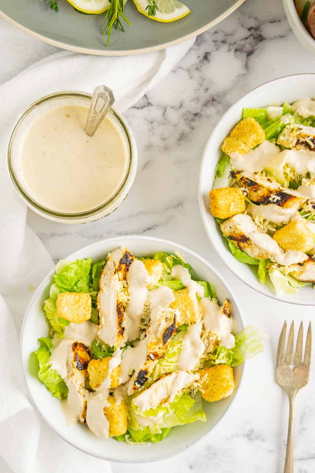 A jar of caesar dressing with a spoon in it and bowls of chicken caesar salad.
