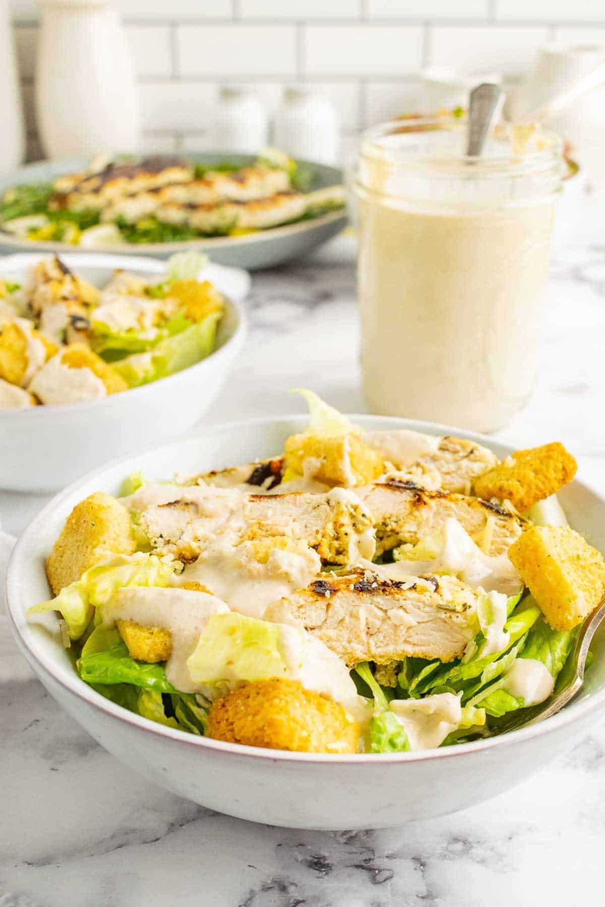 A white bowl with chicken caesar salad and a jar of dressing behind it.