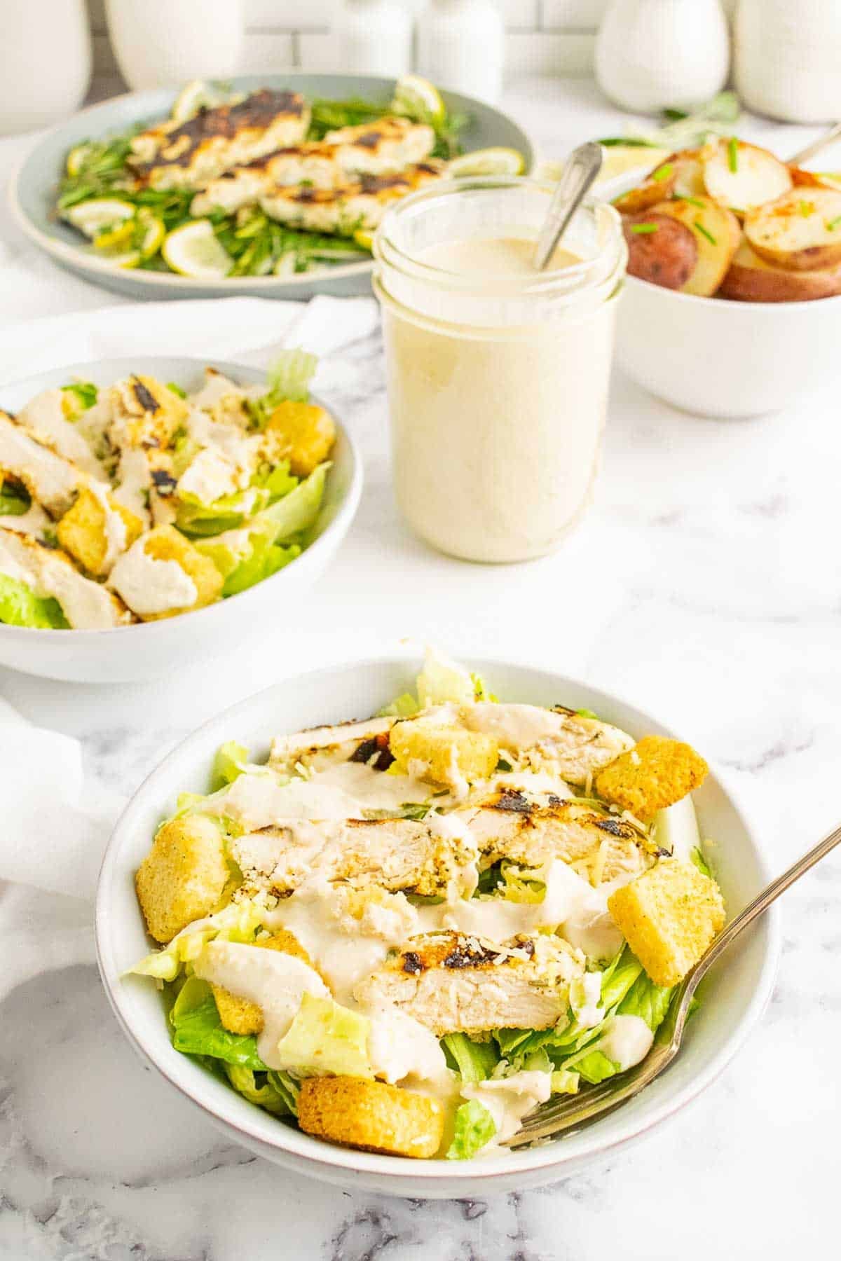 Bowls of chicken caesar salad with a jar of dressing in the middle.