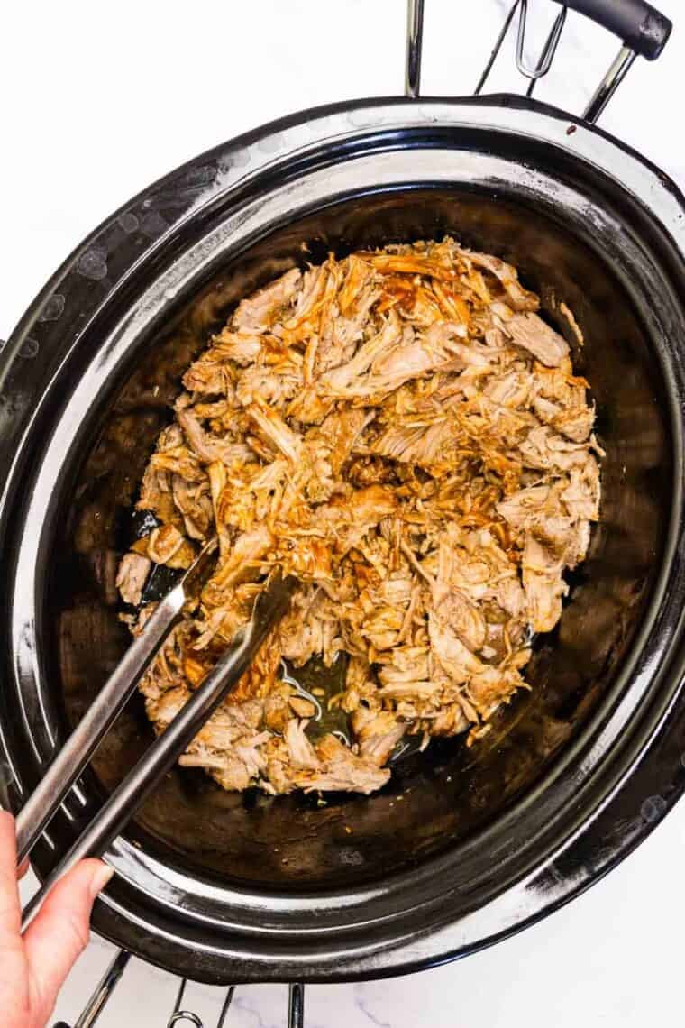 Tongs mix in sauce to a slow cooker of pulled pork.