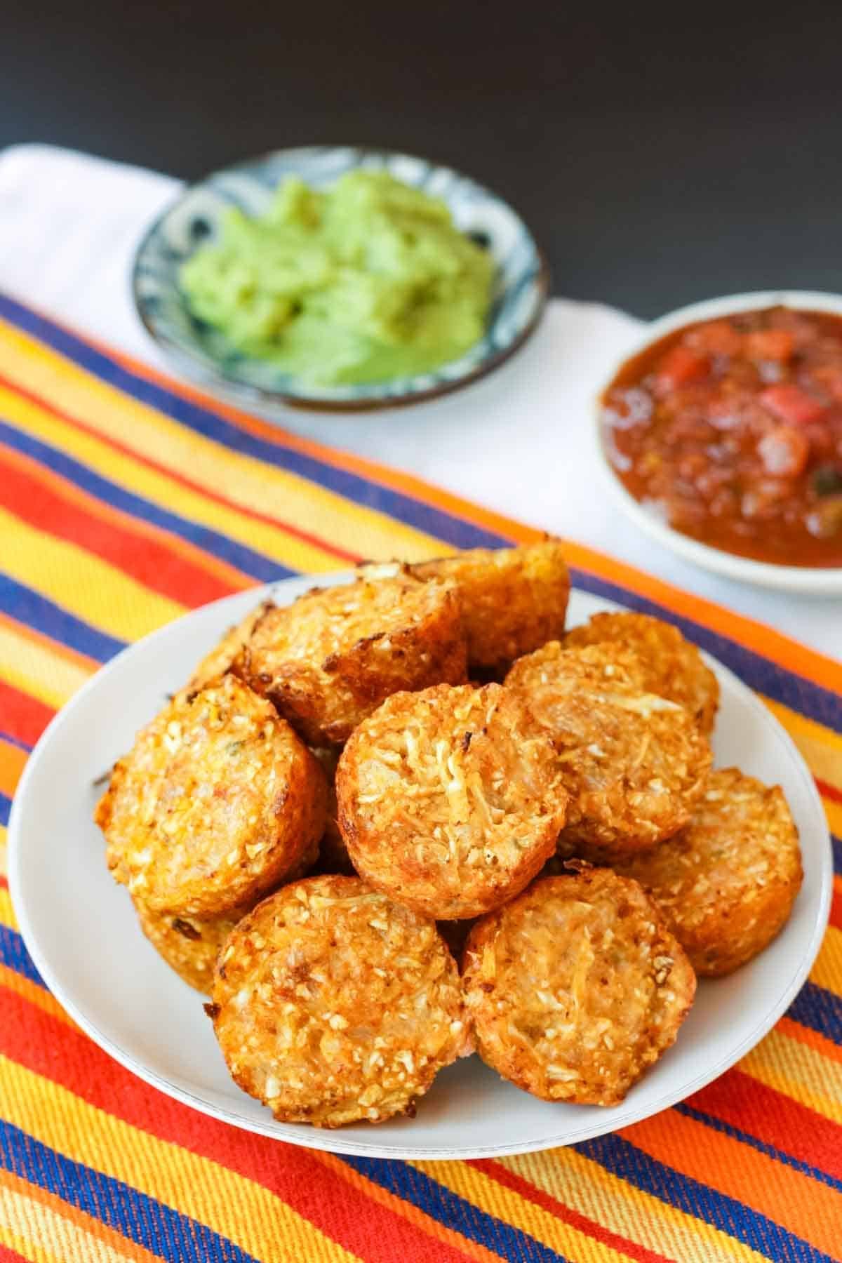 A plateful of cauli tots on a striped napkin with small dishes of guacamole and salsa behind it.