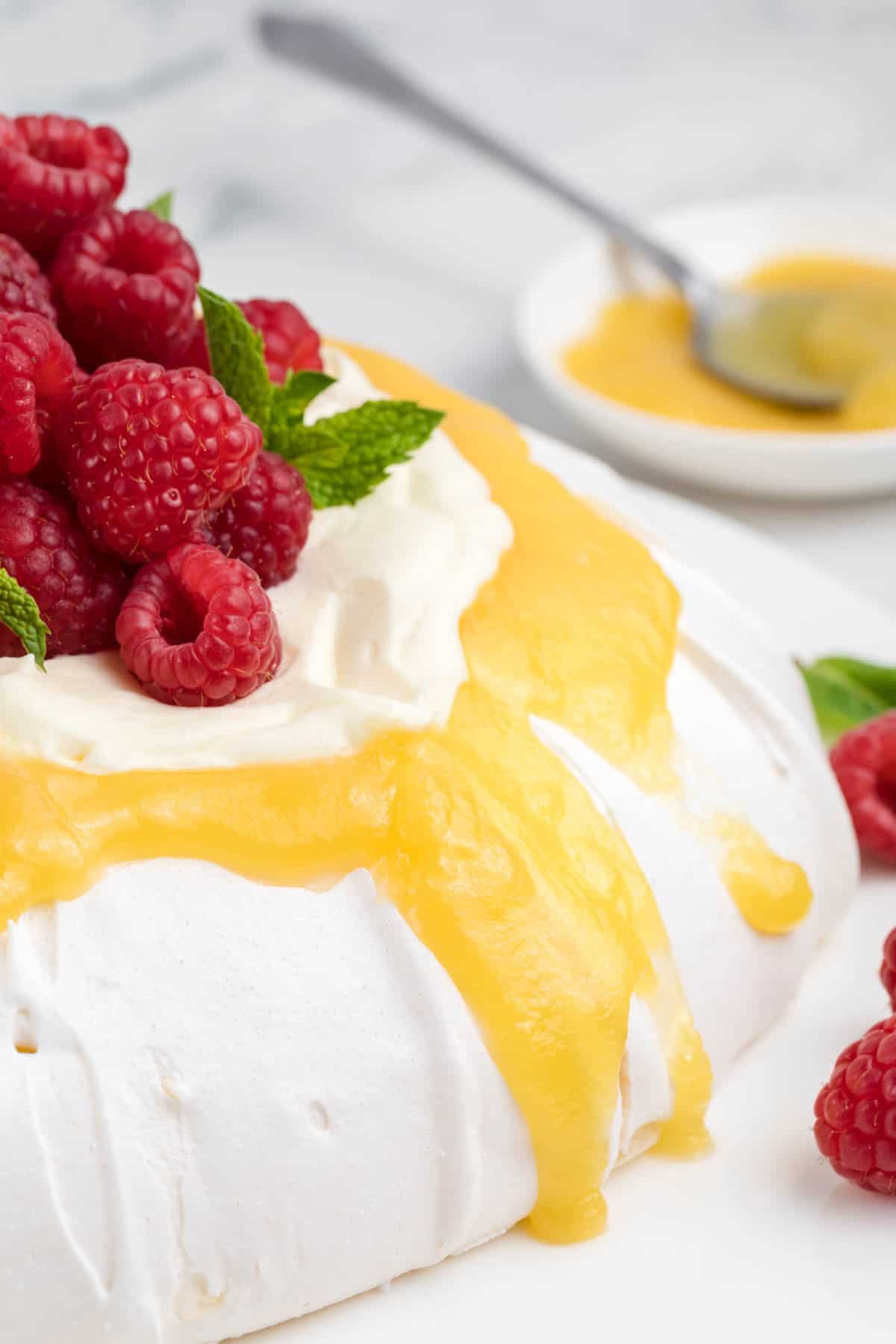 Lemon curd drips down the sides of a pavlova that's also topped with whipped cream and raspberries.