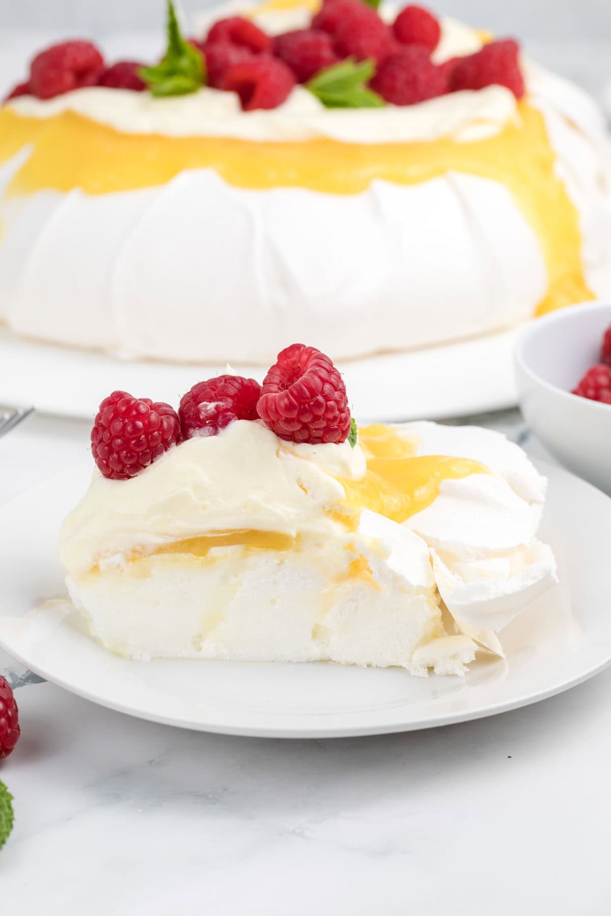 A slice of pavlova topped with lemon curd, whipped cream, and raspberries.