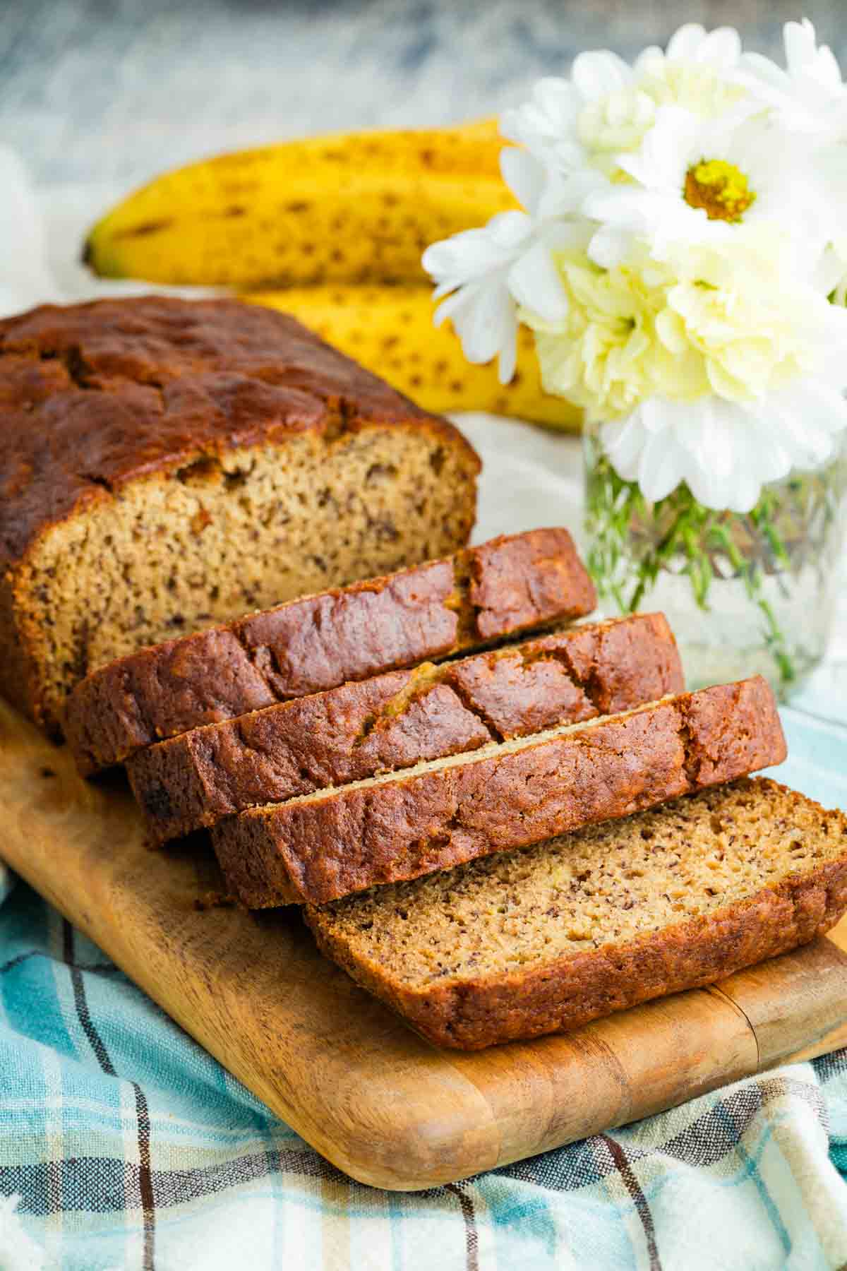 A fresh loaf of gluten-free banana bread cut into slices with bananas and flowers nearby.