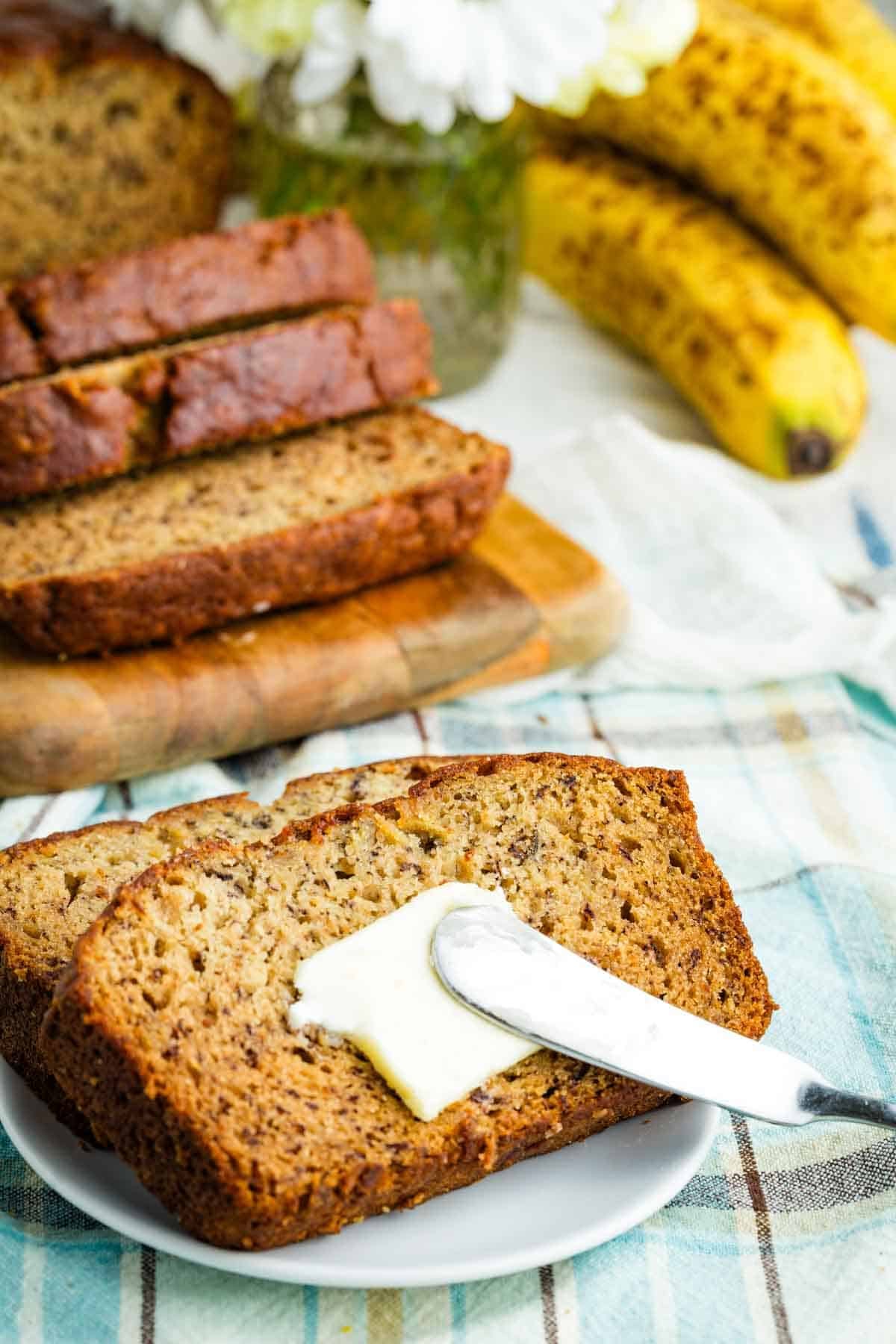A knife spreads butter on a slice of gluten-free banana bread.
