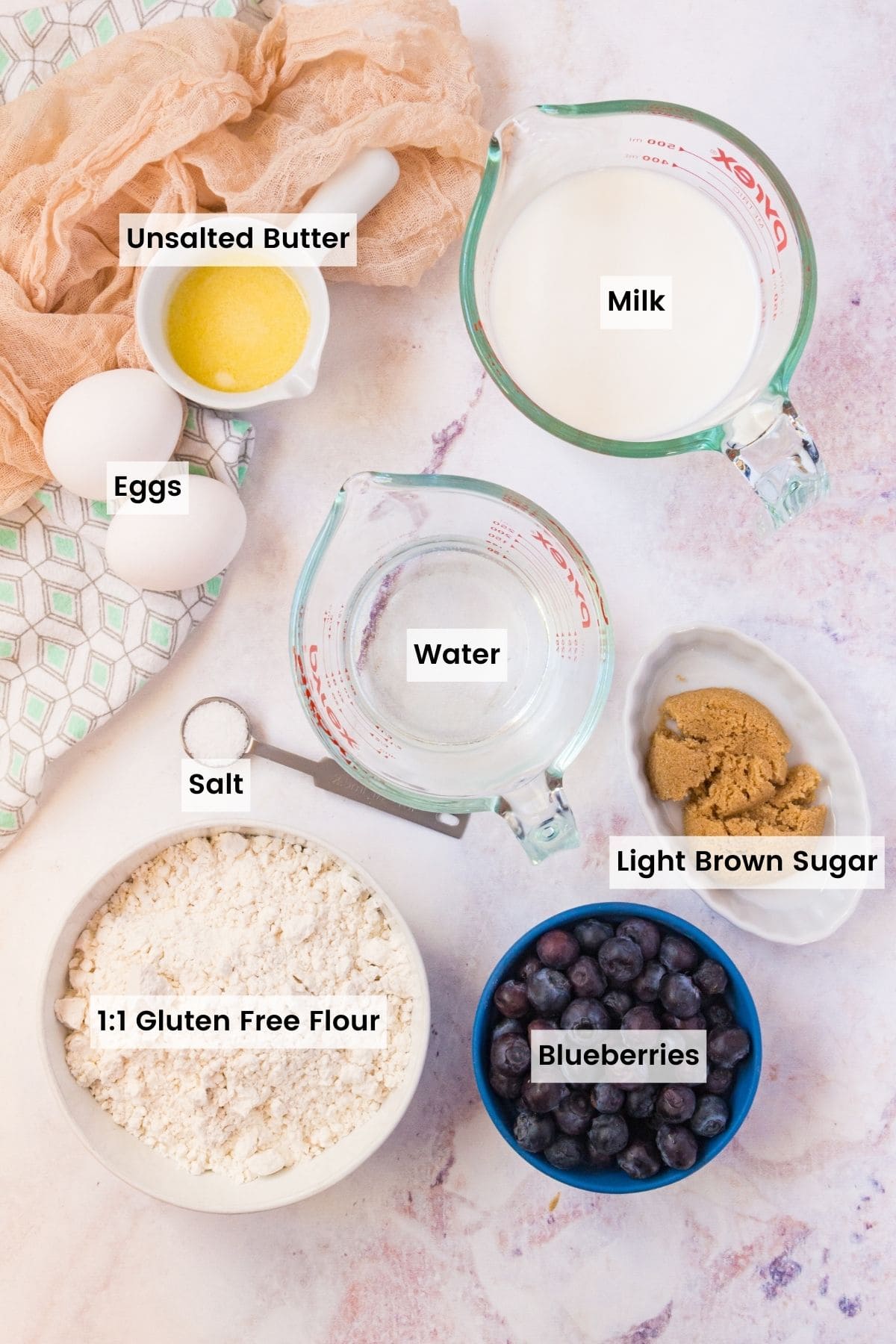 Milk, blueberries, brown sugar, eggs and the rest of the ingredients on a countertop with text labels.