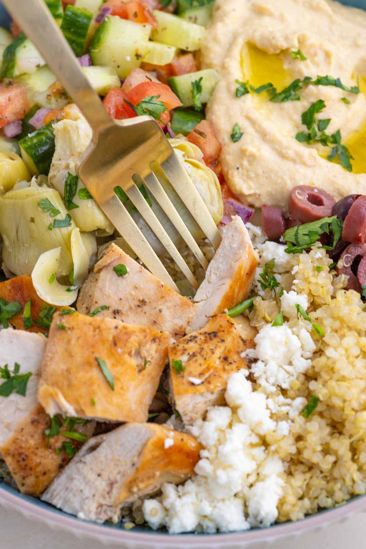 These easy Greek Chicken Bowls are loaded with fresh veggies, kalamata olives, and artichoke hearts served over quinoa and topped with hummus. Closeup image of a fork diving into a Greek chicken bowl.