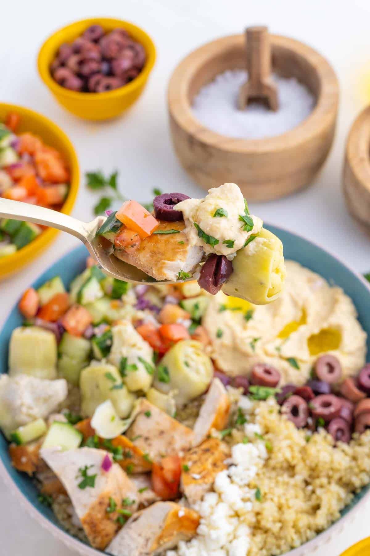 These easy Greek Chicken Bowls are loaded with fresh veggies, kalamata olives, and artichoke hearts served over quinoa and topped with hummus. A fork taking a bite from a Greek chicken bowl.