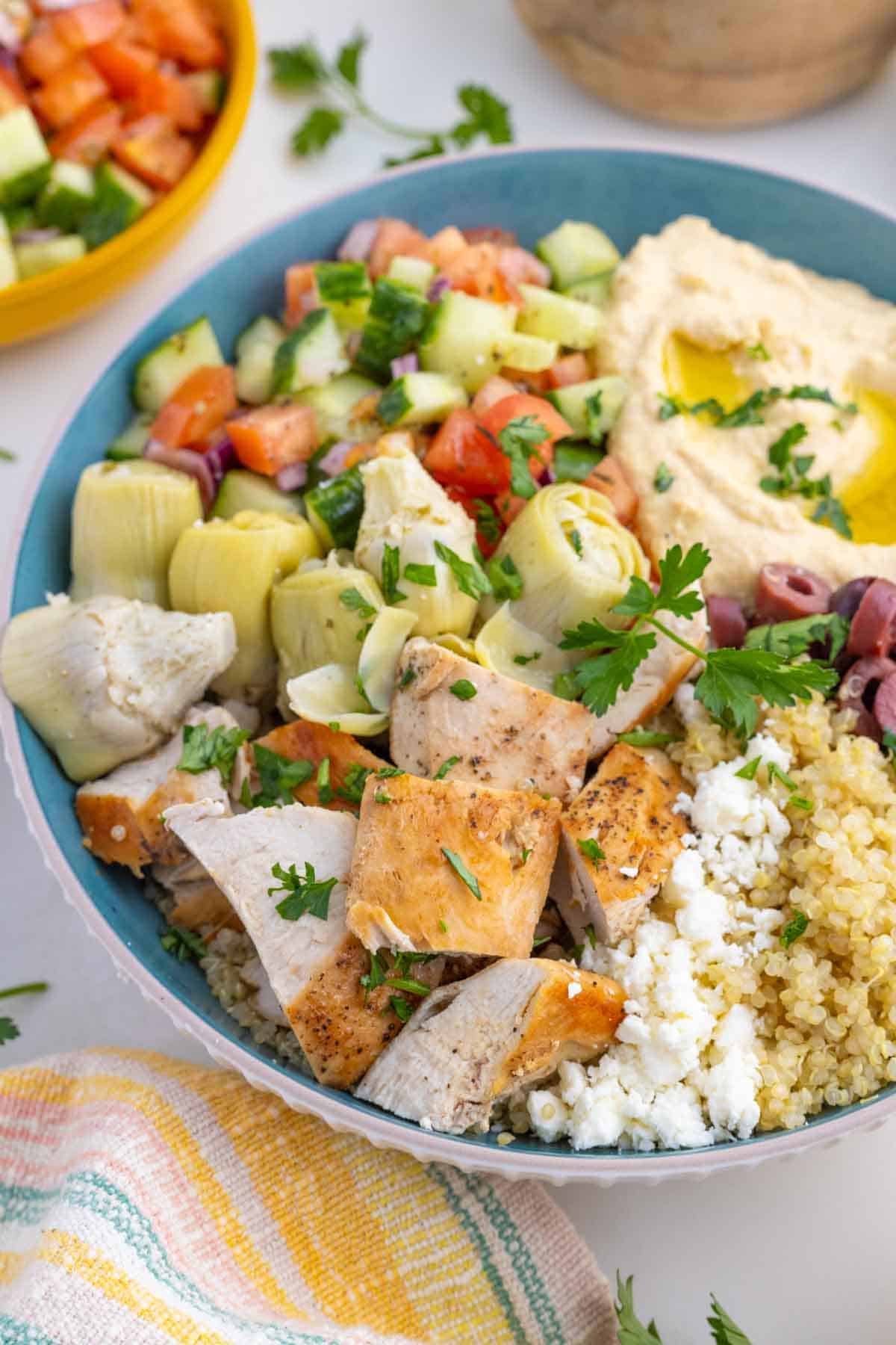 These easy Greek Chicken Bowls are loaded with fresh veggies, kalamata olives, and artichoke hearts served over quinoa and topped with hummus. Close-up image of a Greek chicken bowl with hummus.