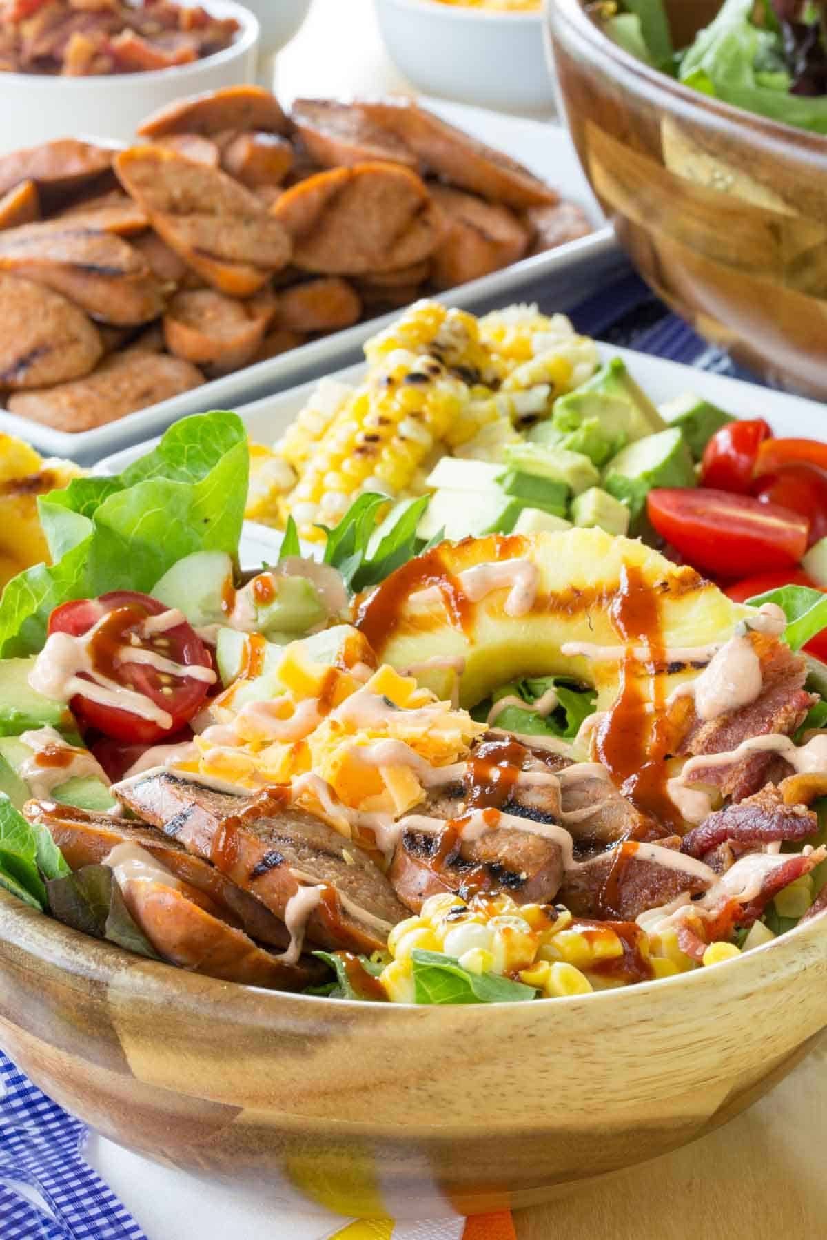 Grilled chicken sausage salad features a mix of chicken sausage, pineapple, fresh corn, avocado, and lettuce, drizzled with creamy dressing. A bowl of chicken sausage salad topped with hot sauce.