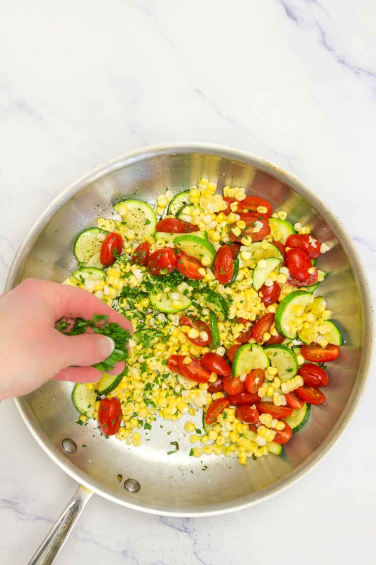 A hand adds herbs to a pan of satueed vegetables.