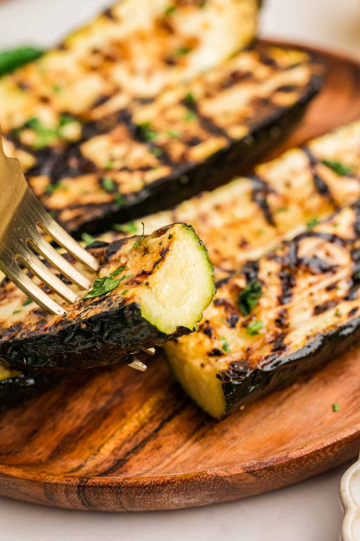 A fork taking a bite of grilled zucchini on a wooden plate.