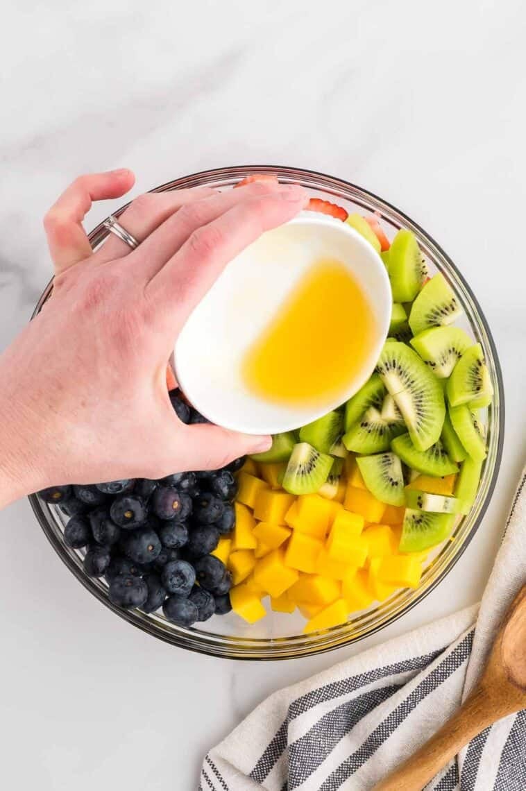 Pouring honey-lime dressing over blueberries, mangos, kiwi, and strawberries in a bowl.