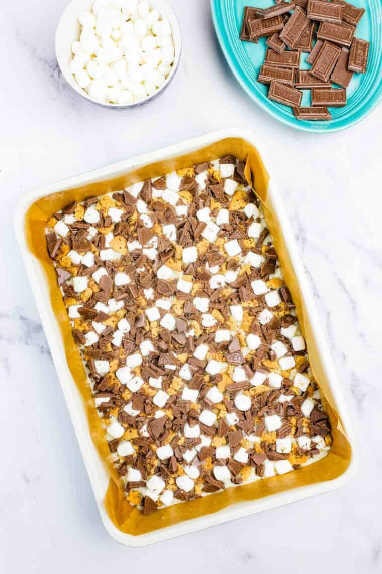 Scattering chocolate pieces, mini marshmallows, and graham cracker pieces over a graham cracker crust covered in sweetened condensed milk.