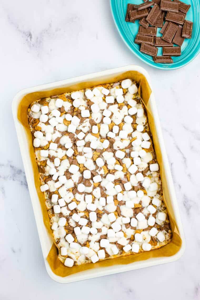 Scattering extra mini marshmallows over a graham cracker crust covered in sweetened condensed milk, chocolate pieces, graham cracker pieces, and more mini marshmallows.