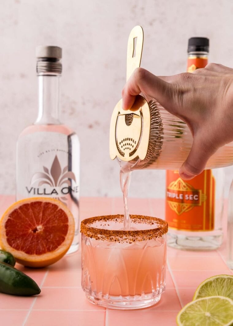 Pouring a grapefruit margarita from a cocktail shaker into a glass.