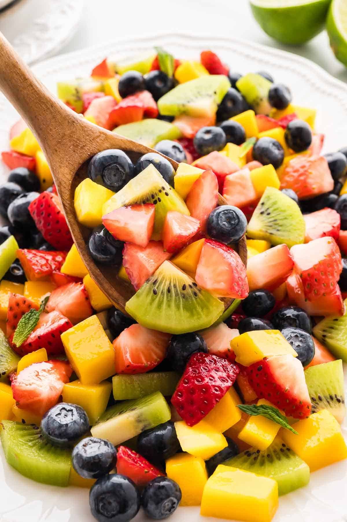 This Mango Berry Fruit Salad is a refreshing summer treat, featuring sweet, seasonal fruit and a tangy honey-lime dressing. A wooden spoon in mango berry fruit salad.