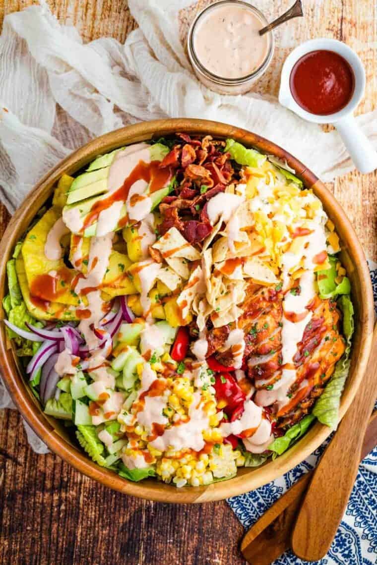 This BBQ chicken salad is loaded with BBQ chicken, crispy bacon, juicy pineapple, creamy avocado, and more. A sweet and savory masterpiece. Overhead image of BBQ chicken and pineapple salad in a wooden salad bowl (before tossing).