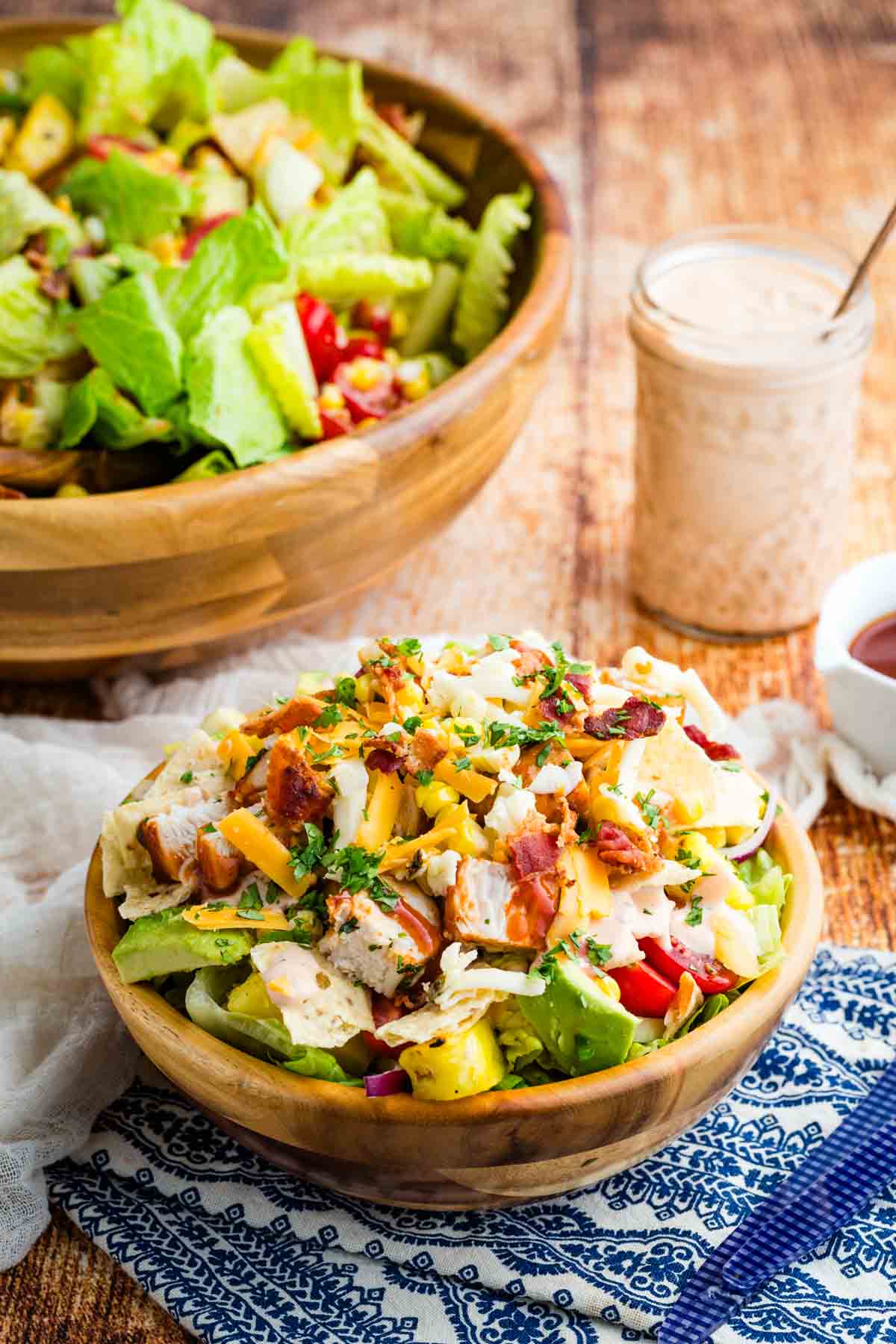 This BBQ chicken salad is loaded with BBQ chicken, crispy bacon, juicy pineapple, creamy avocado, and more. A sweet and savory masterpiece. BBQ chicken salad in a wooden bowl with a lager bowl of salad in the background.