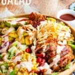 A salad topped with chicken, corn, bacon, cheese, tomatoes, and avocado in a large wood bowl with text overlay that says "BBQ Chicken Salad".