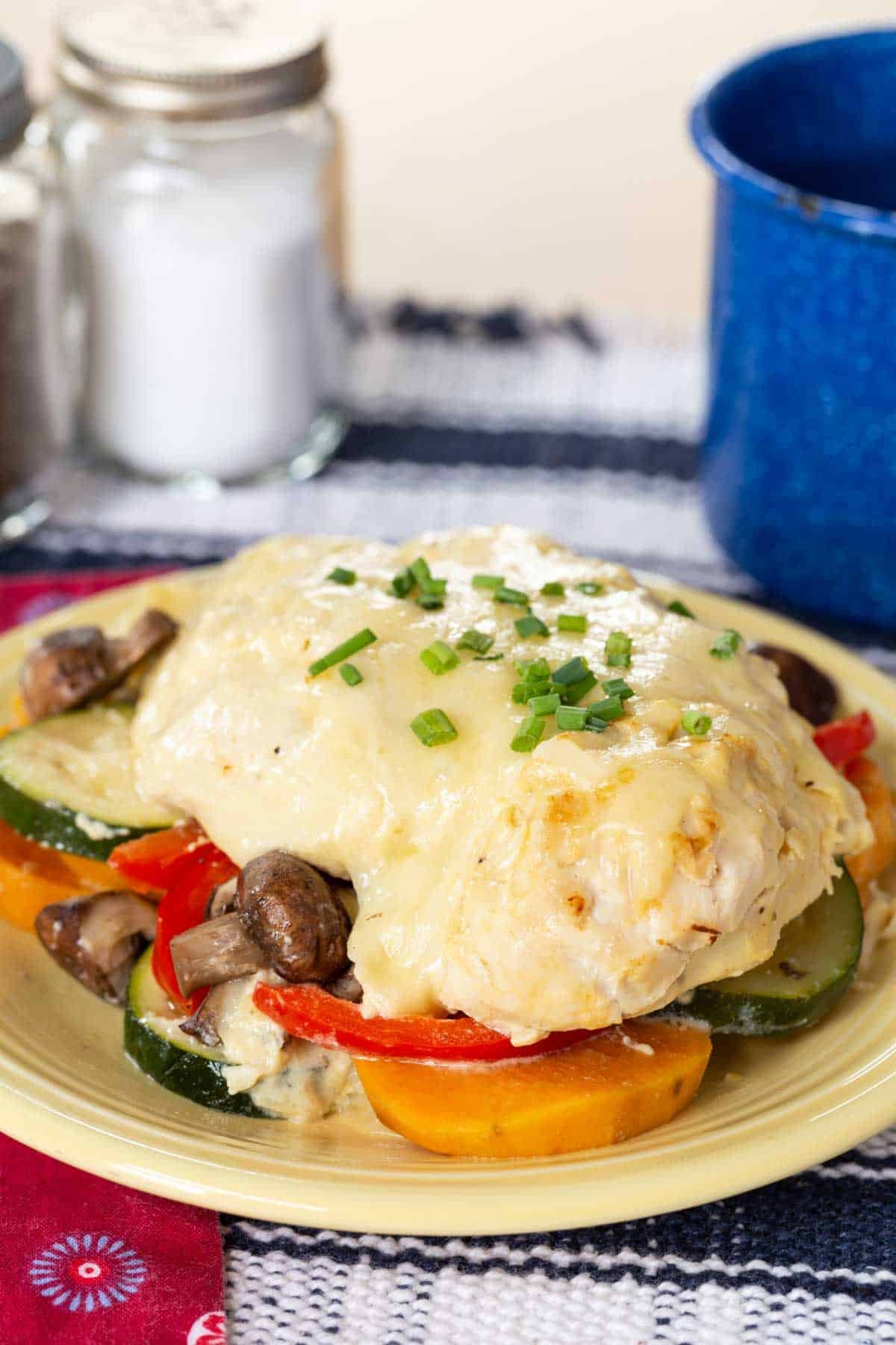 Chicken topped with cheese resting on a mixture of veggies on a yellow plate.