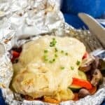 Chicken and veggies in a foil pouch on a blue plate with text overlay that says "Cheesy Chicken Foil Packets".