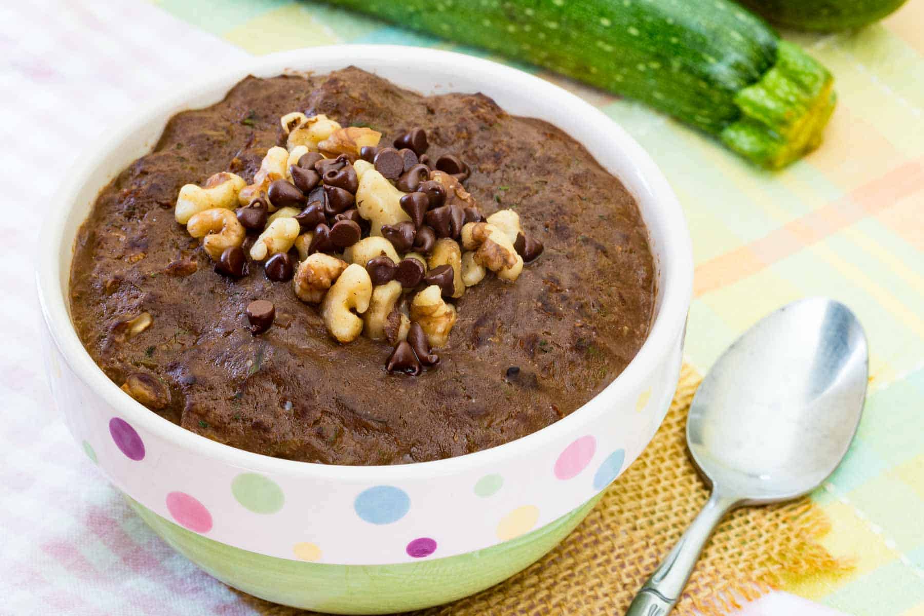 A bowl of chocolate oatmeal topped with walnuts and chips with zucchini in the background.
