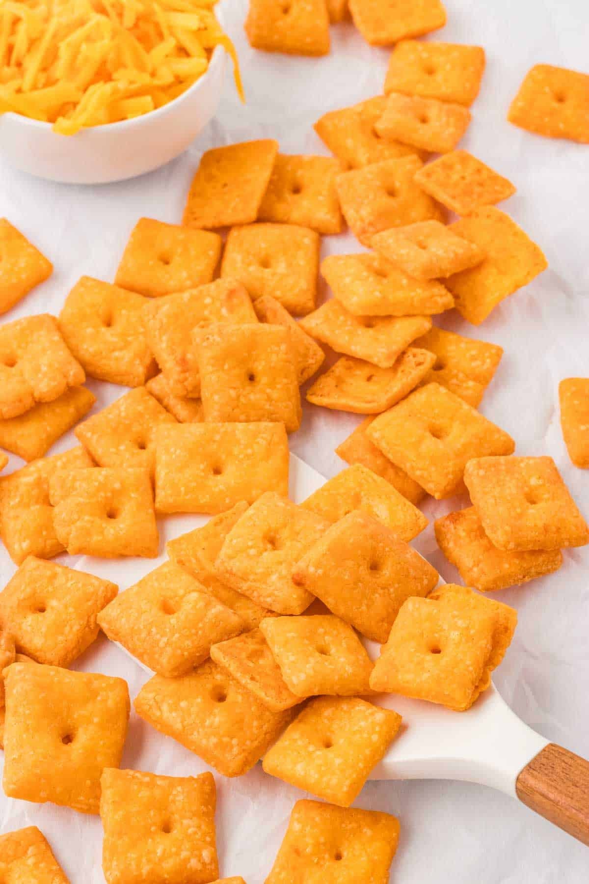 A rubber spatula scooping through cheddar crackers piled onto parchment paper.