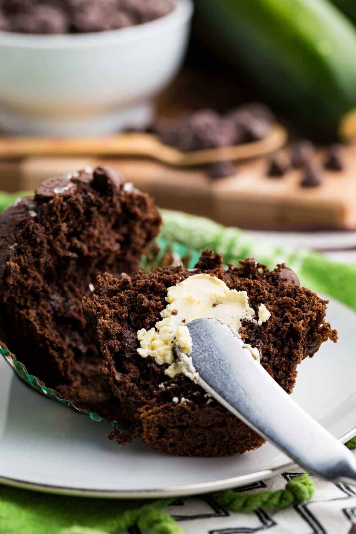 These gluten-free chocolate zucchini muffins are packed with melty chocolate chips and incredibly moist and tender, thanks to the zucchini. Buttering a gluten free chocolate zucchini muffin.