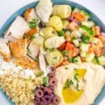 A bowl of quinoa topped with chicken, hummus, veggies, olives, and feta cheese with text overlay that says "Greek Chicken Bowls".
