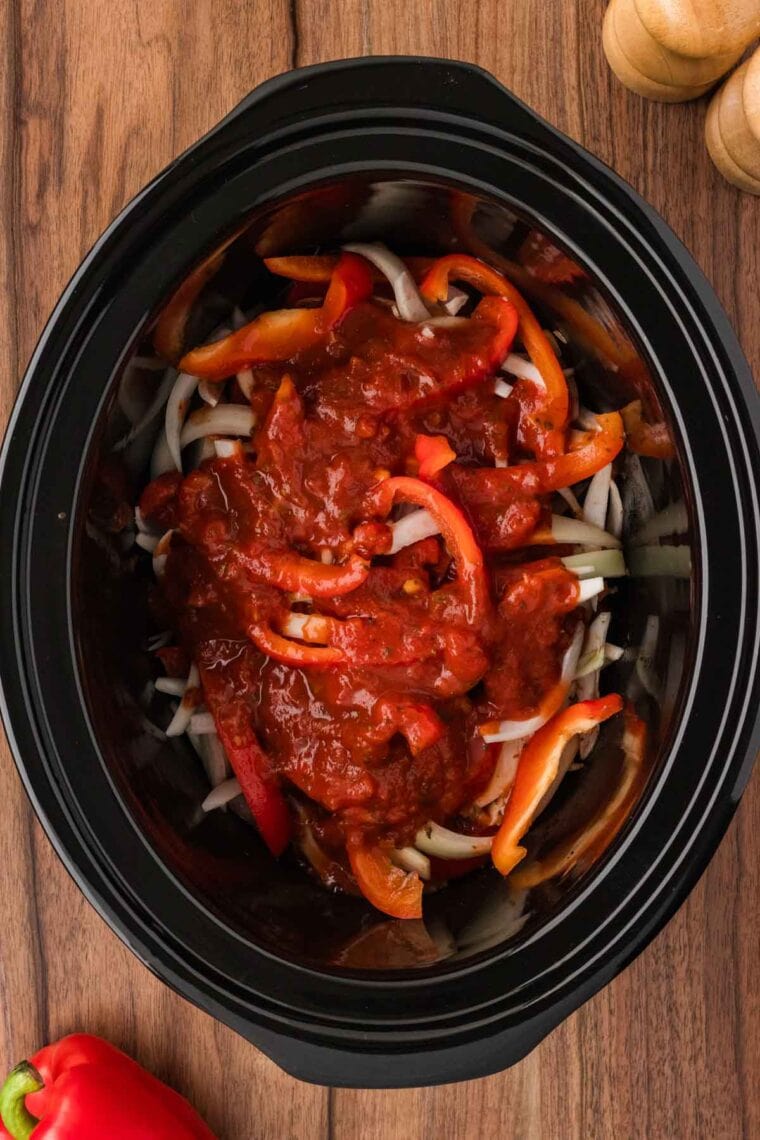 Pouring tomato sauce over a seasoned London broil, bell peppers, and onions in a slow cooker.