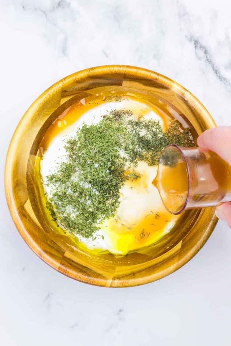 This Greek yogurt ranch dip is full of bold, herby flavors, perfect for scooping up with dippables or spreading it over a juicy burger. Pouring apple cider vinegar into a bowl with Greek yogurt, herbs, onion powder, garlic powder, salt, pepper, and olive oil.