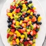 A platter of chopped strawberries, kiwi, mango, and blueberries with text overlay that says "Mango Berry Fruit Salad".
