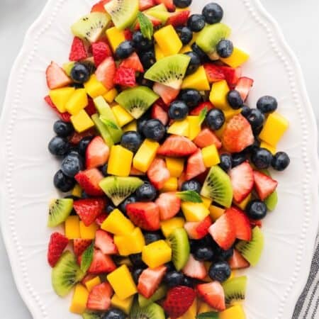A platter of chopped strawberries, kiwi, mango, and blueberries with text overlay that says "Mango Berry Fruit Salad".