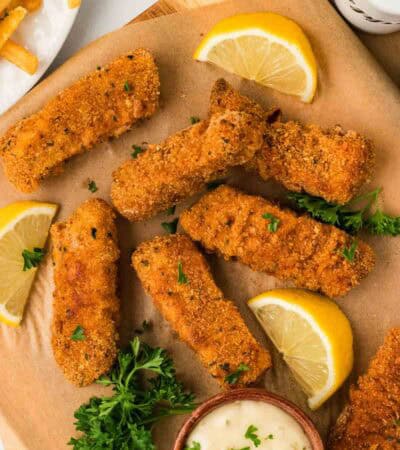 Gluten free fish sticks on parchment paper with tartar sauce and lemon wedges.
